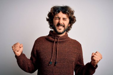 Young handsome man with beard wearing casual sweater standing over white background very happy and excited doing winner gesture with arms raised, smiling and screaming for success. Celebration concept.