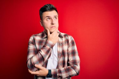 Kırmızı izole edilmiş arka plan üzerine gündelik modern tişört giyen yakışıklı beyaz bir adam bir soru hakkında endişeli ve gergin bir şekilde düşünüyor.