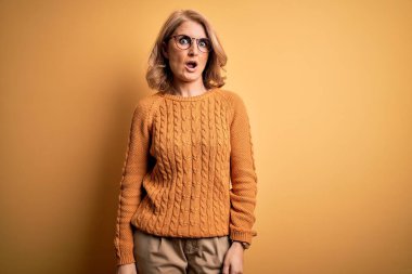 Orta yaşlı, sarışın bir kadın sarı arka planda günlük süveter ve gözlük takıyor. Şok suratlı, şüpheci ve alaycı görünüyor.