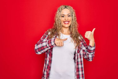 Genç, sarışın, gündelik bir gömlek giyen, kırmızı arka planda duran, el ve baş parmaklarıyla arkasını gösteren, kendinden emin bir şekilde gülümseyen bir kadın.