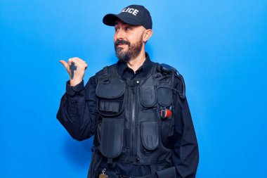 Polis üniforması giyen, gülümseyen, gülümseyen ve baş parmağıyla yan tarafı gösteren genç yakışıklı adam.. 