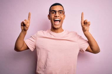 Yakışıklı Afro-Amerikalı adam pembe arka planda günlük tişört ve gözlük takıyor. Gülümsüyor, şaşırıyor, parmaklarıyla işaret ediyor ve kollarını kaldırıyor..