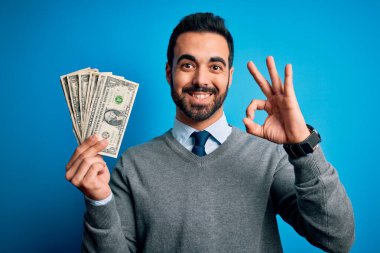 Sakallı, mavi arka planda banknotları olan yakışıklı bir genç. Tamam işareti yapıyor, mükemmel bir sembol.