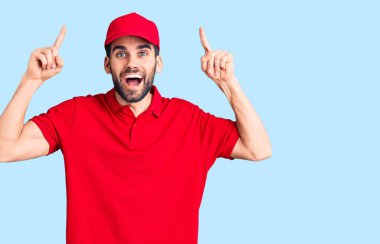 Sakallı yakışıklı adam teslimat üniforması giyiyor. Şaşırmış, şaşırmış, parmaklarıyla işaret ediyor ve kollarını kaldırıyor.. 