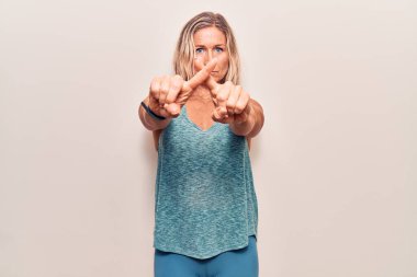 Orta yaşlı, beyaz, spor kıyafetleri giyen sarışın bir kadın. Eksi işareti yaparken şans diliyor. 