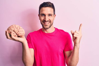 Genç ve yakışıklı bir adam beyni soyutlanmış pembe arka planda tutmak istiyor. Gülümsüyor, elini ve parmağını yana uzatarak işaret ediyor.