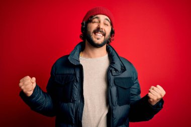 Sakallı, sıradan bir ceket giyen, kırmızı arka planda yün şapka takan çok mutlu ve heyecanlı genç bir adam. Kolları havada, gülümseyen ve başarı için çığlık atan, kazanan bir jest yapıyor. Kutlama konsepti.