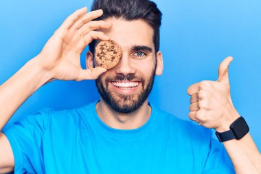 Sakallı, elinde çikolatalı kurabiyeyle mutlu ve pozitif gülümseyen genç ve yakışıklı bir adam, başparmağıyla mükemmel ve onay işareti yapıyor. 