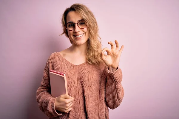 Fiatal Gyönyörű Szőke Visel Szemüveg Gazdaság Notebook Felett Rózsaszín Háttér — Stock Fotó