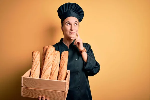 Young beautiful baker woman wearing cooker uniform and hat holding wooden box with bread serious face thinking about question, very confused idea