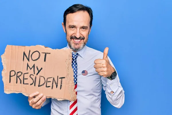 Middle age business man asking for political holding banner with not my president message smiling happy and positive, thumb up doing excellent and approval sign