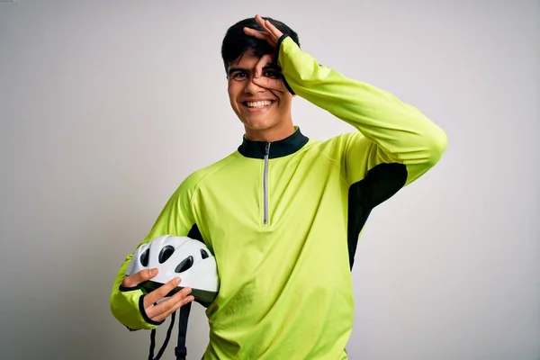 Young handsome cyclist man holding security bike helmet over isolated white background with happy face smiling doing ok sign with hand on eye looking through fingers