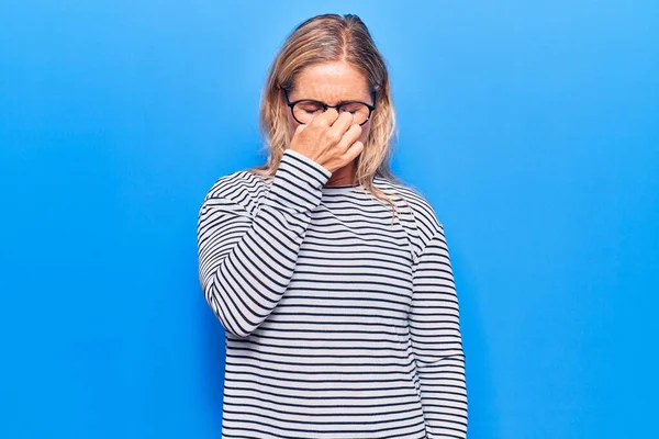 Middle age caucasian blonde woman wearing casual striped sweater and glasses tired rubbing nose and eyes feeling fatigue and headache. stress and frustration concept.