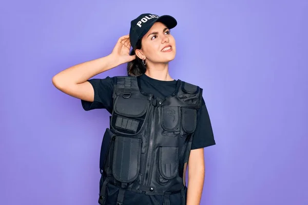 Young police woman wearing security bulletproof vest uniform over purple background smiling confident touching hair with hand up gesture, posing attractive and fashionable