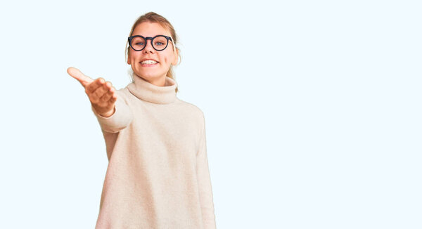 Young beautiful blonde woman wearing turtleneck sweater and glasses smiling friendly offering handshake as greeting and welcoming. successful business. 