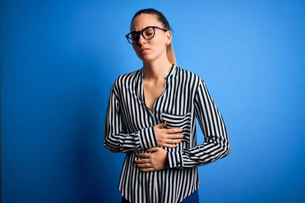 Beautiful blonde woman with blue eyes wearing striped shirt and glasses over blue background with hand on stomach because indigestion, painful illness feeling unwell. Ache concept.