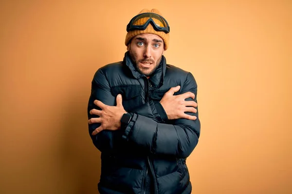 Young handsome skier man with beard wearing snow sportswear and ski goggles shaking and freezing for winter cold with sad and shock expression on face