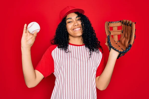 Young african american curly sportswoman wearing cap playing baseball using ball and glove with a happy face standing and smiling with a confident smile showing teeth