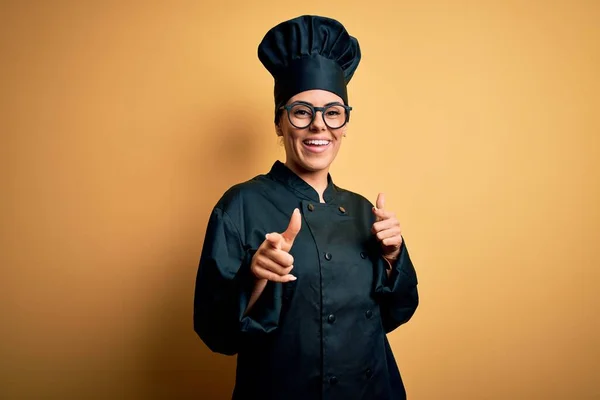 Young beautiful brunette chef woman wearing cooker uniform and hat over yellow background pointing fingers to camera with happy and funny face. Good energy and vibes.