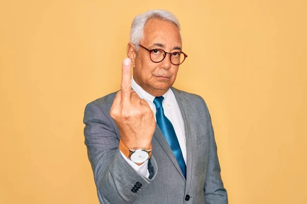 Middle age senior grey-haired handsome business man wearing glasses over yellow background Showing middle finger, impolite and rude fuck off expression