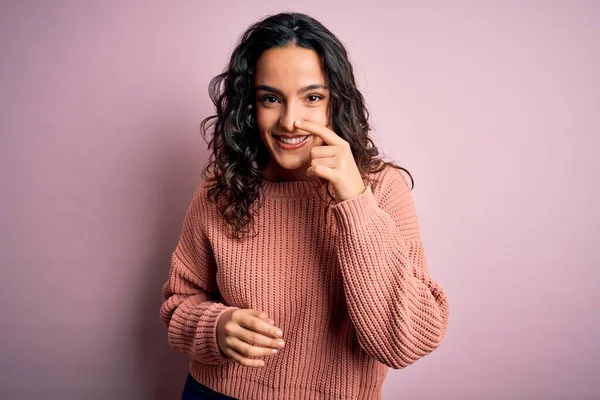 Young beautiful woman with curly hair wearing casual sweater over isolated pink background Pointing with hand finger to face and nose, smiling cheerful. Beauty concept