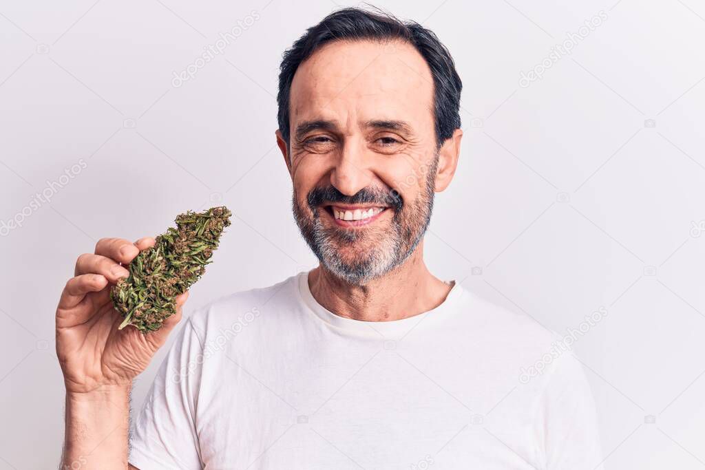 Middle age handsome man holding cannabis weed standing over isolated white background looking positive and happy standing and smiling with a confident smile showing teeth