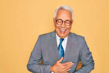 Orta yaşlı, gri saçlı, yakışıklı iş adamı sarı arka planda gözlük takıyor, gülümsüyor ve kahkahalarla gülüyor..