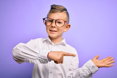 Mor arka planda mavi gözlü, beyaz gömlekli, beyaz gözlü küçük beyaz çocuk kameraya hayret ediyor ve gülümsüyor..