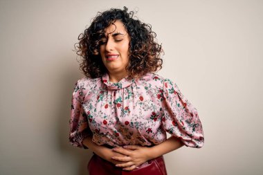 Genç ve güzel kıvırcık Arap kadın çiçekli tişört giyer. İzole edilmiş beyaz arka plan üzerinde durur. Midenin üstünde el ele durur çünkü hazımsızlık, acı veren bir hastalık kendini iyi hissetmez. Ache kavramı.