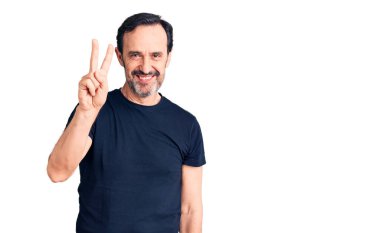 Orta yaşlı yakışıklı bir adam sıradan tişörtler giyer ve parmaklarıyla iki numarayı gösterirken kendinden emin ve mutlu bir şekilde gülümser.. 