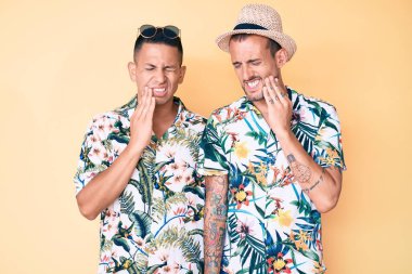 İki genç eşcinsel erkek yaz şapkası takıyor ve Hawaii gömleği diş ağrısı ya da diş ağrısı yüzünden ağızlarına acı dolu bir ifadeyle dokunuyor. dişçi 