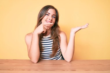 Masada oturan, elleri ve parmaklarıyla kameraya gülümseyen, gündelik giysiler giyen güzel, beyaz bir kadın.. 