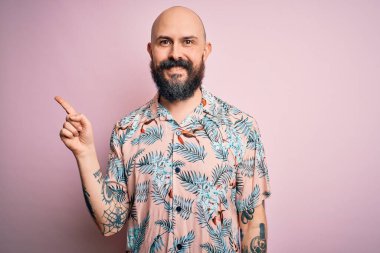 Sakallı ve dövmeli yakışıklı, pembe arka planda sıradan bir çiçek gömleği giyen, yüzünde kocaman bir gülümseme olan, eli kameraya bakan kel bir adam..