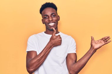 Genç Afro-Amerikalı, günlük kıyafetler giymiş, avuç içi gösteriyor ve baş parmağıyla iyi bir hareket yapıyor, mutlu ve neşeli gülümsüyor. 