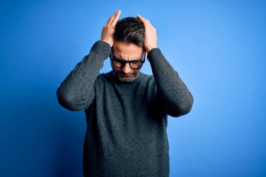 Genç, yakışıklı bir adam, günlük süveter ve gözlük takıyor mavi arka planda dikiliyor. Ağrı ve migren yüzünden baş ağrısı çekiyor ve stresli. Ellerini başının üstüne koy..