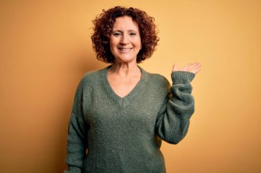 Orta yaşlı, kıvırcık saçlı güzel bir kadın izole edilmiş sarı arka planda günlük süveter giyiyor neşeli bir şekilde gülümsüyor ve avuç içi kameraya bakıyor..