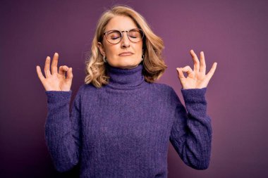 Orta yaşlı, sarışın, sıradan mor balıkçı yaka kazak giyen, gözlüklü, gözleri kapalı gülümseyen, meditasyon yaparken parmaklarıyla gülümseyen güzel bir kadın. Yoga konsepti.