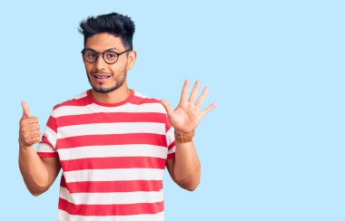 Yakışıklı Latin Amerikalı genç adam gündelik kıyafetler ve gözlük takıyor. Altıncı parmaklarını gösterip gösterirken kendinden emin ve mutlu gülümsüyor.. 