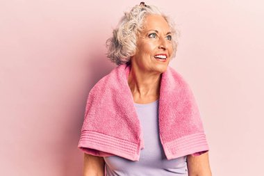 Spor kıyafetleri ve havlu giyen yaşlı, gri saçlı bir kadın yan tarafa bakıyor, rahat profil pozu veriyor doğal bir yüz ve kendine güvenen bir gülümsemeyle.. 