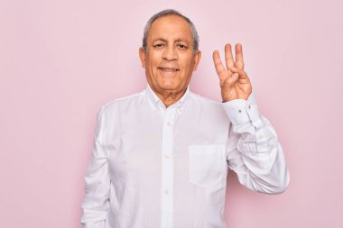 Üst düzey yakışıklı, gri saçlı, zarif tişörtlü, soyutlanmış pembe arka planda, üç numarayı işaret eden, kendinden emin ve mutlu gülümseyen bir adam..