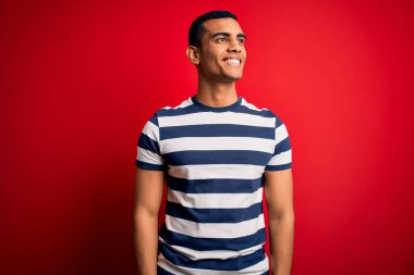 Yakışıklı Afro-Amerikan erkeği, sıradan çizgili tişört giyiyor kırmızı arka planda dikiliyor yüzünde gülümseme, doğal bir ifade ile yan yana bakıyor. Kendinden emin gülen.