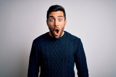 Sakallı, sıradan bir süveter giyen genç ve yakışıklı bir adam beyaz arka plan üzerinde korku dolu ve şaşkınlık dolu bir ifade ve heyecanlı bir yüz ifadesiyle duruyor..
