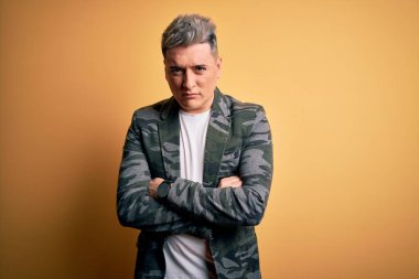 Sarı izole edilmiş arka plan üzerinde duran yakışıklı, modern bir adam şüpheci ve gergin, yüzünde çapraz kollu tasvip etmeyen bir ifade. Negatif kişi.