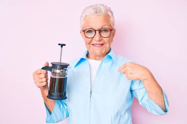 Mavi gözlü, gri saçlı, Fransız kahve makinesini tutan ve gülümseyen mutlu ve gururlu bir kadını işaret eden son sınıf güzeli. 