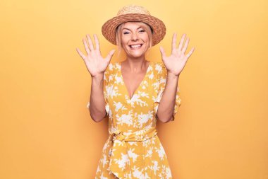Sarışın, güzel bir kadın tatilde. Sarı arka planda şapkası ve elbisesi var. Onuncu parmaklarıyla işaret ediyor, kendinden emin ve mutlu gülümsüyor..