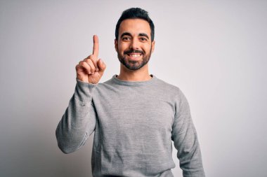 Sakallı, sıradan bir süveter giyen, beyaz arka plan üzerinde duran ve bir numaralı parmağını gösterip gülümseyen, kendinden emin ve mutlu genç bir adam..