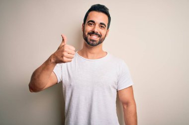 Sakallı, sıradan tişörtlü, beyaz arka planda duran genç ve yakışıklı bir adam elinle mutlu bir jest yapıyor. Kameraya bakıp başarıyı gösteren ifade onaylanıyor.