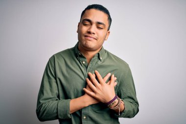 İzole edilmiş beyaz arka plan üzerinde dikilen Brezilyalı genç adam elleri göğsünde, gözleri kapalı ve yüzünde minnet dolu bir jest ile gülümsüyor. Sağlık kavramı.