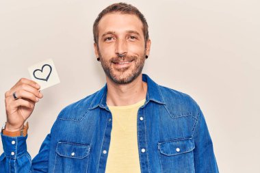 Elinde kalp anımsatıcı tutan genç yakışıklı. Olumlu ve mutlu görünüyor. Kendine güvenen bir gülümsemeyle ayakta duruyor ve dişlerini gösteriyor. 