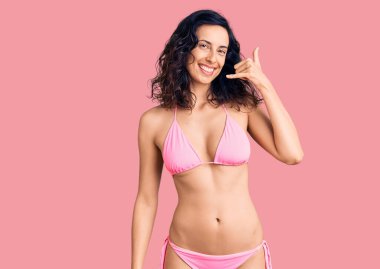 Bikini giyen, gülümseyen, el ve parmaklarıyla telefon hareketi yapan, telefonda konuşur gibi genç ve güzel bir İspanyol kadın. iletişim kavramları. 